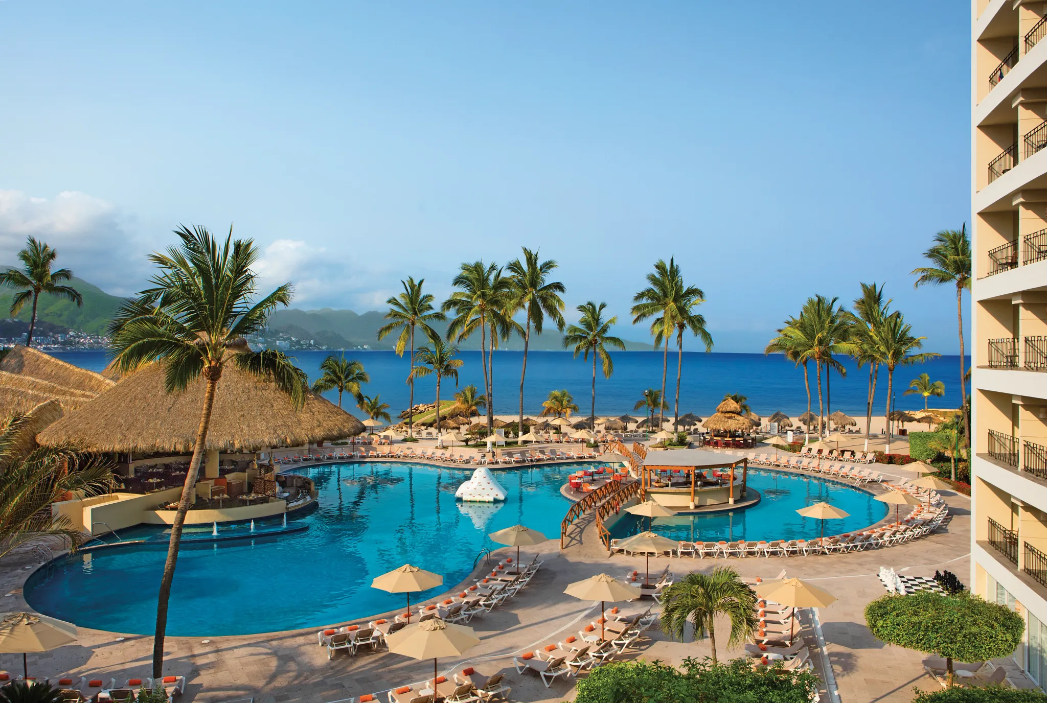 Panoramic view of Sunscape Puerto Vallarta Resort & Spa pools with palm trees, ocean backdrop, and beachfront lounge areas.